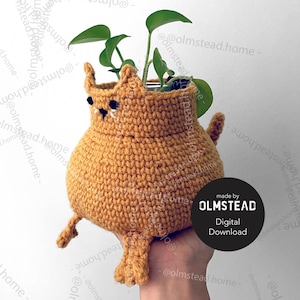 May include: A crocheted cat planter in a light brown color with a small green plant inside. The planter has a cute face with black eyes and a small black nose. The planter is being held by a hand.