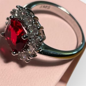 Peut inclure: Une bague en argent avec une grande pierre précieuse rouge carrée entourée de petites pierres précieuses claires. La bague a une bande simple.