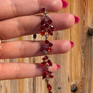 May include: A silver chain with dangling red crystal drops. The chain is made of small, connected links and features a cluster of red crystals at the top.