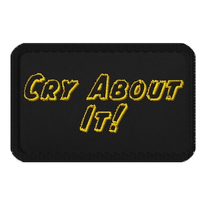 May include: Black rectangular patch with the embroidered phrase "Cry About It!" in yellow, outlined in a darker yellow. The patch has a black border and is likely designed to be sewn onto clothing or accessories.