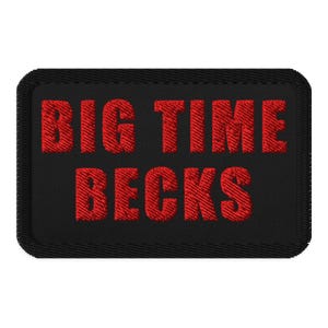 以下が含まれることがあります： 黒い長方形のパッチ。赤い糸で「BIG TIME BECKS」と刺繍されています。テキストは太字で、パッチにはステッチされたボーダーがあります。