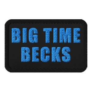 以下が含まれることがあります： 黒い長方形のパッチに、青色で「BIG TIME BECKS」の文字が刺繍されています。テキストは太字のサンセリフ体で、パッチにはステッチされた縁があります。