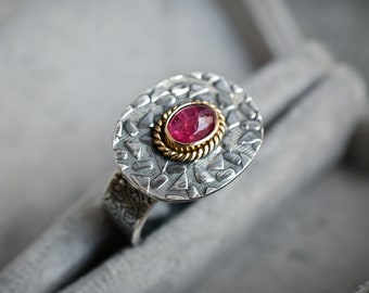 May include: A silver ring with a textured oval band and a pink gemstone set in a gold bezel. The ring is in a gray velvet box.