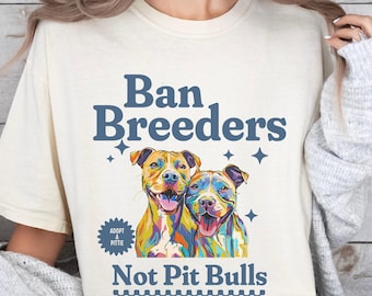 May include: Cream-colored t-shirt with the text "Ban Breeders Not Pit Bulls" in blue. The shirt features a colorful illustration of two smiling pit bull-type dogs. The design includes stars and a small "Adopt a Pittie" badge.