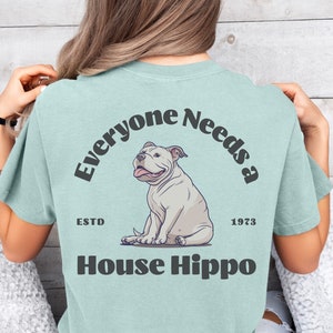 May include: A light blue t-shirt with the text "Everyone Needs a House Hippo" in black. The text is in a semi-circle around a cartoon image of a white bulldog sitting down. The text "ESTD 1973" is below the bulldog.
