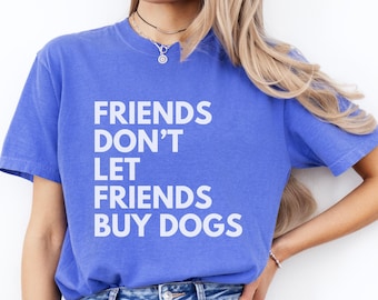 May include: A blue t-shirt with the text "FRIENDS DON'T LET FRIENDS BUY DOGS" in white block letters. The shirt is a solid blue color and has short sleeves. The model is wearing blue jeans.