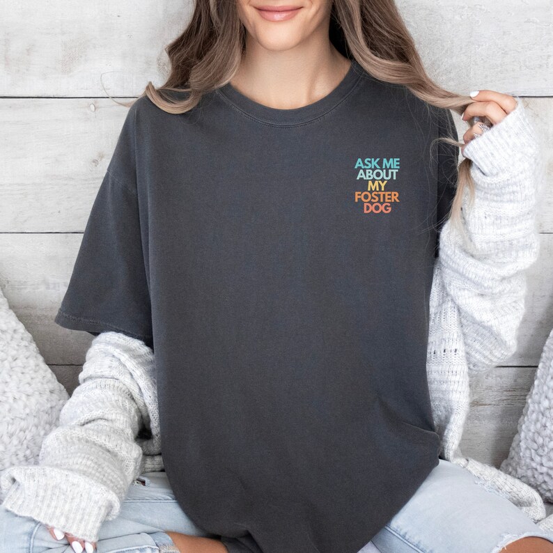 May include: A dark gray t-shirt with the text "ASK ME ABOUT MY FOSTER DOG" printed on the front in a rainbow gradient.