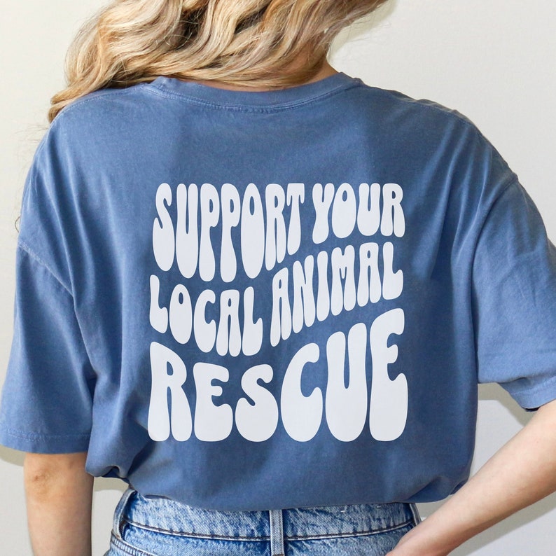 May include: A blue t-shirt with the text "SUPPORT YOUR LOCAL ANIMAL RESCUE" printed in white on the back. The text is in a retro, wavy font.