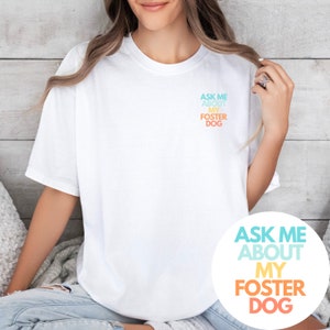 May include: White t-shirt with the text "Ask me about my foster dog" printed on the front in a rainbow gradient. The text is also printed on a white circle.