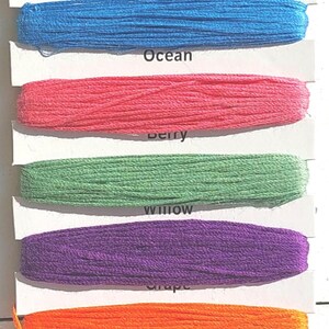May include: A pack of six 8.75 yard spools of 100% cotton embroidery floss in muted colors. The colors are ocean blue, pink, green, purple, orange, and teal. The pack is labeled "Watercolor Muted Scrapbook Stitches".