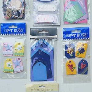 May include: Assortment of paper crafting supplies including collage tags, acrylic labels, tags, and metal accents. The tags are in various colors and designs, including flowers, stars, hearts, and geometric shapes. The metal accents are rectangular frames with a pink, yellow, and blue finish.
