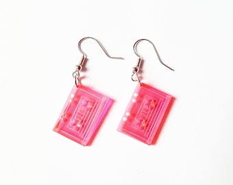Op de afbeelding: Een paar roze cassetteband-oorbellen met zilveren haakjes. De oorbellen zijn gemaakt van helder plastic met een roze tint en hebben een wit cassetteband-ontwerp.