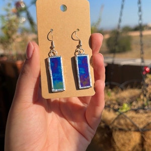 May include: A pair of silver dangle earrings with rectangular blue and purple glass panels. The glass panels have a cloudy, iridescent effect.