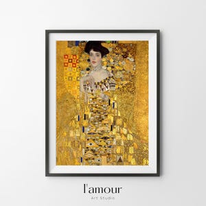 Puede incluir: Impresión enmarcada de una pintura de Gustav Klimt. La obra presenta a una mujer con un vestido dorado y estampado sobre un fondo dorado. La impresión está en un marco negro. El texto "l'amour Art Studio" está en la parte inferior.