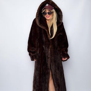 May include: A full-length, dark brown faux fur coat with a large hood. The coat has a button closure and a long, flowing design. The fur appears soft and plush, creating a luxurious look. The coat is worn with a baseball cap and sunglasses.