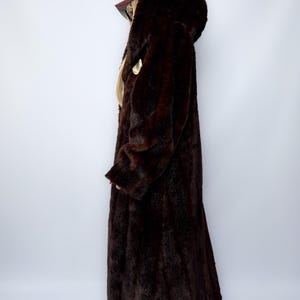 May include: A long, dark brown faux fur coat with a hood. The coat has a full length and a button closure. The coat is a solid color and appears to be made of a soft, plush material. The coat is a classic style.