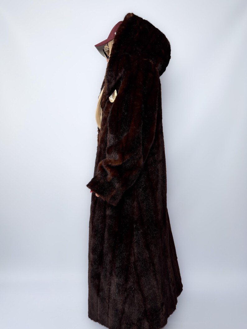 May include: A long, dark brown faux fur coat with a hood. The coat has a full length and a button closure. The coat is a solid color and appears to be made of a soft, plush material. The coat is a classic style.