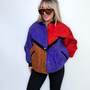 Puede incluir: Chaqueta con bloques de color en tonos morado, rojo y marrón. La chaqueta tiene cierre de botones en la parte delantera y detalles en negro. La chaqueta se lleva con leggings negros. Estilo vintage.