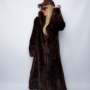 May include: A long, dark brown faux fur coat with a wide collar. The coat is floor-length and has a slight A-line shape. The model is wearing a maroon baseball cap and sunglasses.