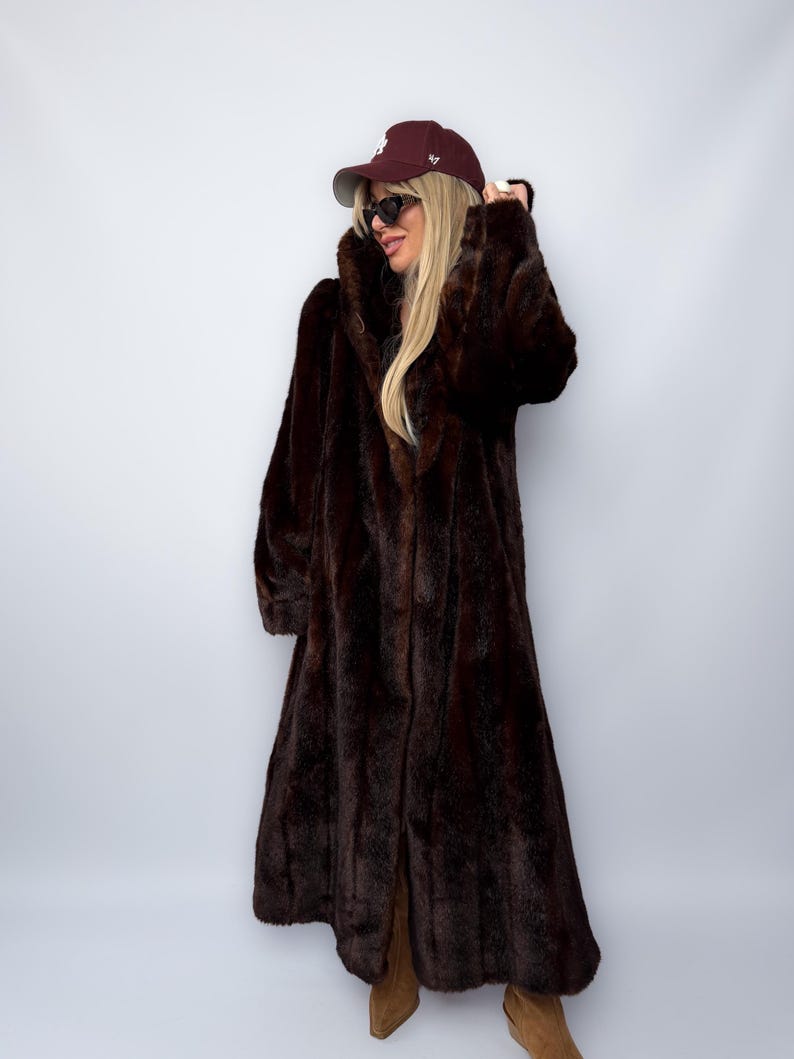 May include: A long, dark brown faux fur coat with a wide collar. The coat is floor-length and has a slight A-line shape. The model is wearing a maroon baseball cap and sunglasses.