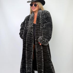 May include: A long, charcoal gray faux fur coat featuring a dark, horizontal tiger-stripe pattern. The coat has a wide shawl collar, long sleeves, and an open front design. It is paired with a black cowboy hat and dark oval-shaped sunglasses.