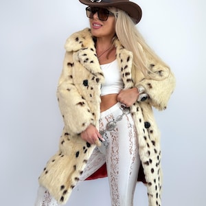 May include: A cream-colored faux fur coat with black spots, paired with white lace pants and a brown cowboy hat. The coat has large black buttons and a red lining. Sunglasses and a silver belt complete the look.