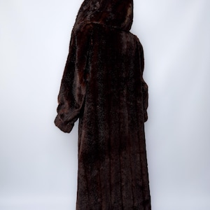 May include: A long, dark brown faux fur coat with a hood. The coat is floor-length and has long sleeves. The person wearing the coat is facing away from the camera, showcasing the coat's texture and design.