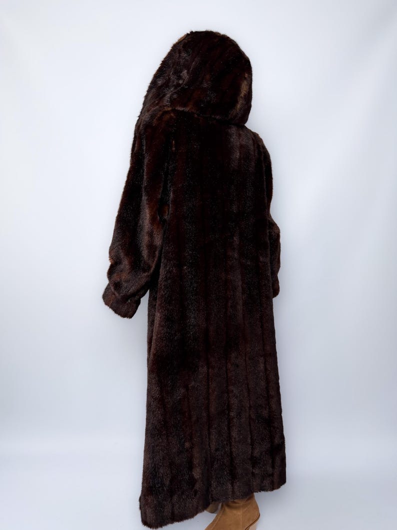 May include: A long, dark brown faux fur coat with a hood. The coat is floor-length and has long sleeves. The person wearing the coat is facing away from the camera, showcasing the coat's texture and design.
