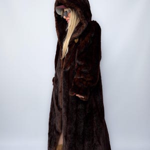 May include: A long, dark brown faux fur coat with a large hood. The coat has a button closure and a full-length design. The model is wearing sunglasses and a cap. The coat is a vintage style.