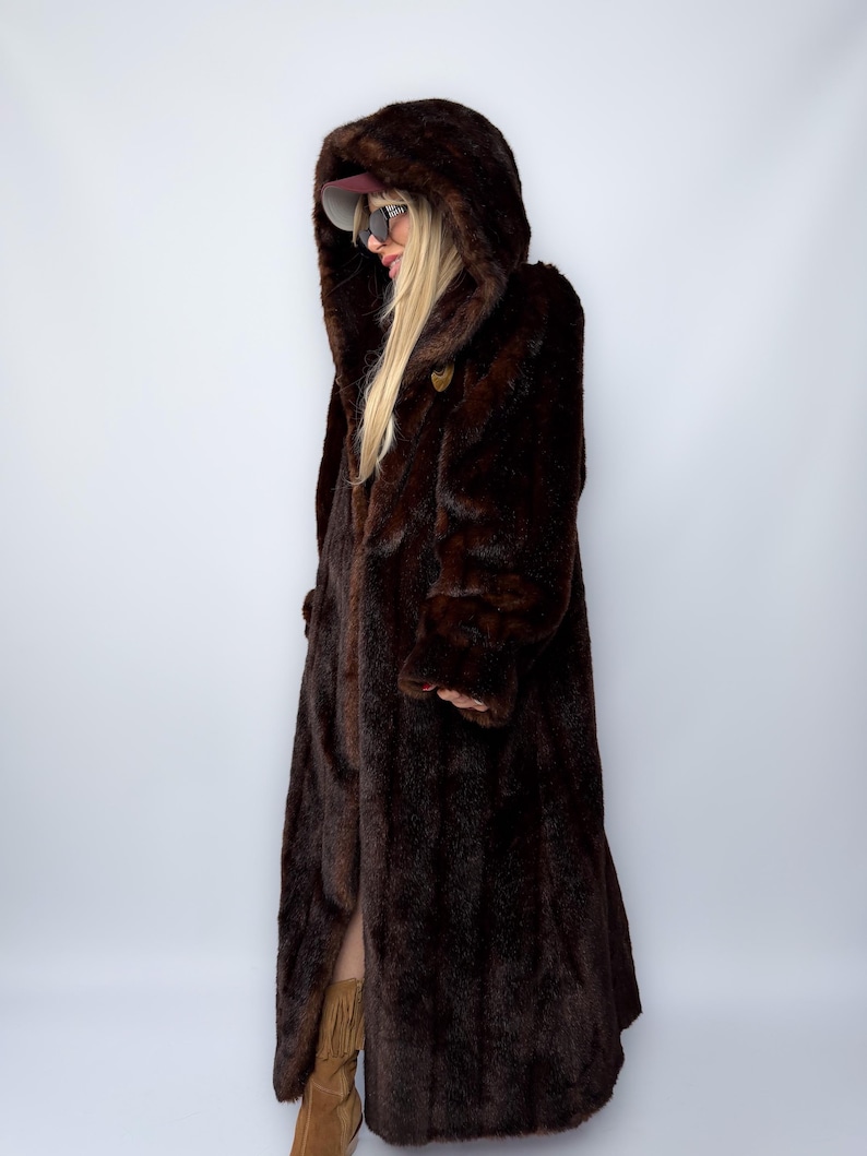 May include: A long, dark brown faux fur coat with a large hood. The coat has a button closure and a full-length design. The model is wearing sunglasses and a cap. The coat is a vintage style.