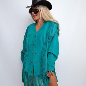 May include: A vibrant teal suede jacket featuring long fringe detailing along the shoulders and front panels. The jacket has a V-neckline, long sleeves, and is fastened down the center with a series of small, circular, metallic decorative buttons.