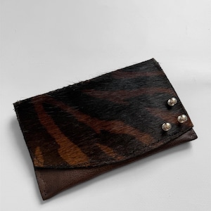 May include: A rectangular wallet with a brown and black zebra stripe pattern on the top flap. The bottom flap is a solid brown leather. Three silver rivets secure the top flap.