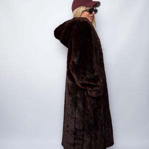 May include: A long, dark brown faux fur coat with a hood. The coat is worn with a maroon baseball cap and sunglasses. The coat appears to be floor-length and has long sleeves. The overall style is vintage and glamorous.