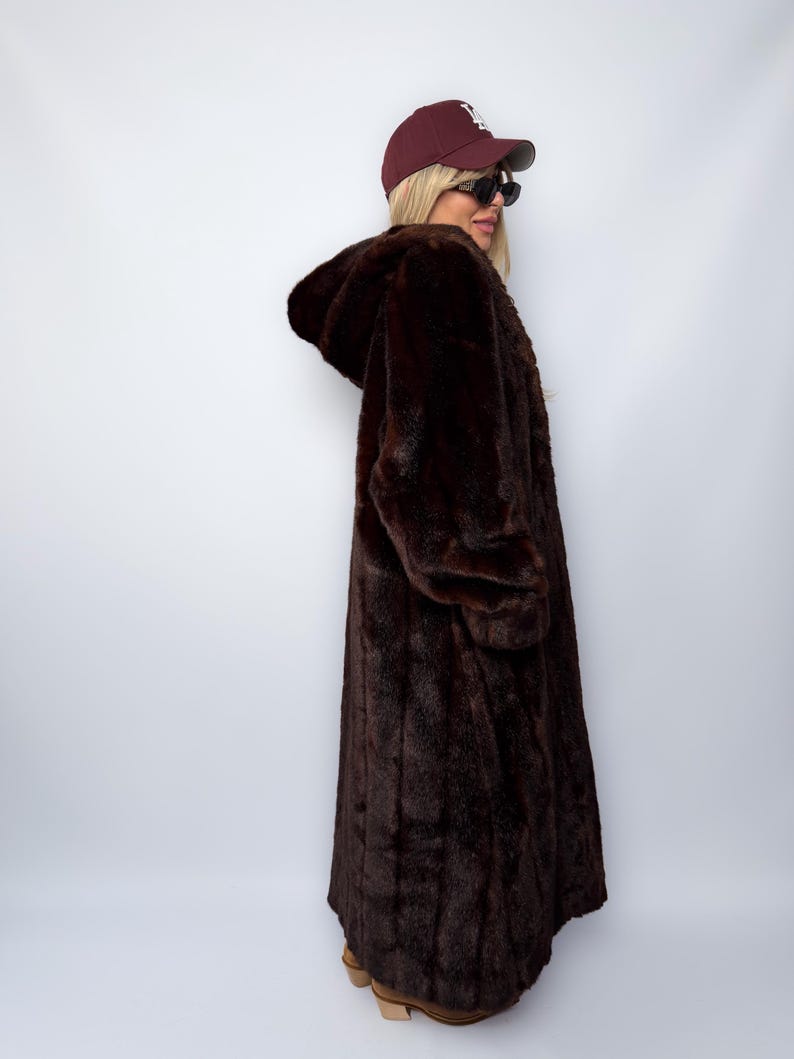 May include: A long, dark brown faux fur coat with a hood. The coat is worn with a maroon baseball cap and sunglasses. The coat appears to be floor-length and has long sleeves. The overall style is vintage and glamorous.