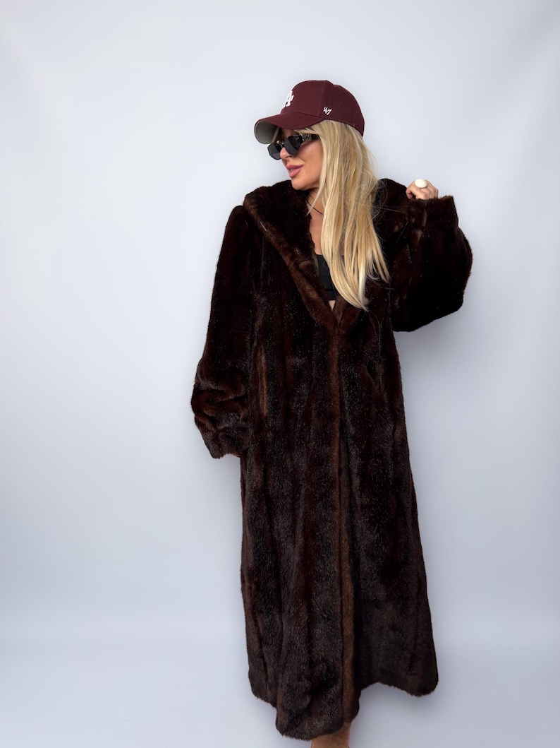 May include: A long, dark brown faux fur coat with a hooded design. The coat is floor-length and has a plush, textured appearance. A maroon baseball cap and sunglasses are also visible.