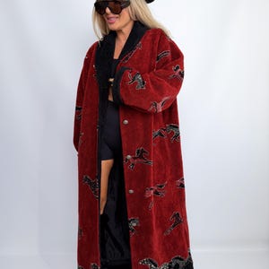May include: A long, deep red coat featuring a repeating pattern of black and white horses. The coat has a black collar, black trim along the hem, and silver-toned metal buttons down the front. The fabric has a textured, plush appearance.