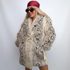 May include: A long-sleeved faux fur coat featuring a cream base color with dark brown and black spotted patterns. The coat has a wide lapel collar, a straight hemline, and a visible white retail tag attached to the lower left side of the garment.