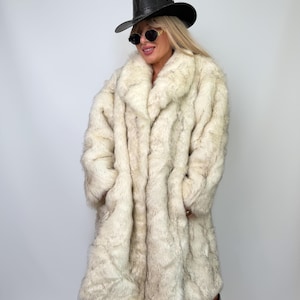 Può includere: Un lungo cappotto in pelliccia sintetica color crema con fibre dalle punte marrone scuro, caratterizzato da un ampio colletto e maniche lunghe. Il cappotto è abbinato a un cappello nero a tesa larga, occhiali da sole rotondi scuri e stivali neri.