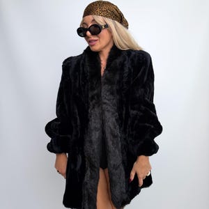 May include: A black faux fur coat with long sleeves and a wide collar, paired with a leopard print headscarf, black oval-framed sunglasses with gold accents, and tan suede knee-high boots against a plain white background.