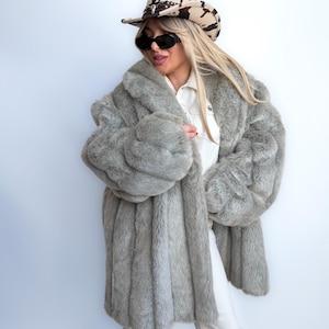 May include: A light gray faux fur coat with a wide collar and long sleeves. The coat is worn with a patterned cowboy hat, sunglasses, white shirt, and pants. Brown fringed boots complete the look. The coat has a vertical ribbed design.