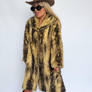Può includere: Un lungo cappotto in pelliccia sintetica con un motivo maculato a strisce verticali color marrone chiaro e scuro. Il cappotto presenta un ampio colletto e maniche lunghe. L'insieme include un cappello da cowboy in camoscio marrone, occhiali da sole ovali neri e stivali con stampa leopardata.