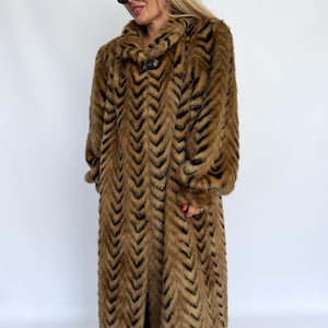 May include: A long, full-length faux fur coat featuring a repeating chevron pattern in shades of brown and black. The coat has a wide collar, long sleeves with gathered cuffs, and a single large dark button closure at the neckline.