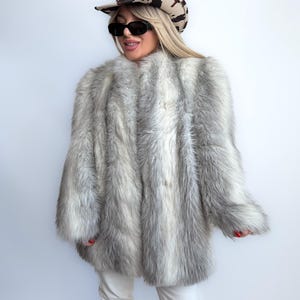 May include: A grey and white faux fur coat with a long, shaggy texture. The coat is worn with a cowboy hat with a cowhide pattern, sunglasses, white trousers, and brown fringed boots. The coat has a classic, oversized shape.