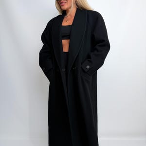 May include: A long, black, open-front coat featuring a wide shawl collar, long sleeves with buttoned cuffs, and a straight-cut silhouette. The coat is worn over a black crop top, paired with black trousers and tan boots, and accessorized with dark sunglasses.