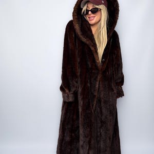 May include: A long, dark brown faux fur coat with a large hood. The coat has a button closure and long sleeves. The hood is up, and the coat appears to be worn over other clothing. The coat is floor-length.