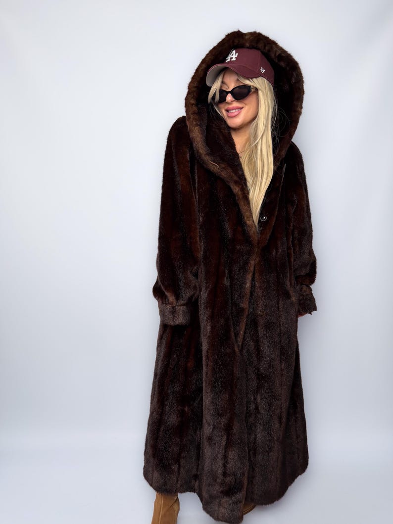 May include: A long, dark brown faux fur coat with a large hood. The coat has a button closure and long sleeves. The hood is up, and the coat appears to be worn over other clothing. The coat is floor-length.