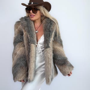 May include: A faux fur coat in shades of grey and tan, with a wide collar and long sleeves. The coat is worn with a brown cowboy hat and sunglasses. The outfit is completed with a white satin dress.