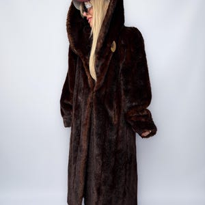 May include: A long, dark brown faux fur coat with a large hood. The coat has a button closure and a soft, plush texture. The hood is lined with a darker shade of brown. A maroon baseball cap is worn on the head.