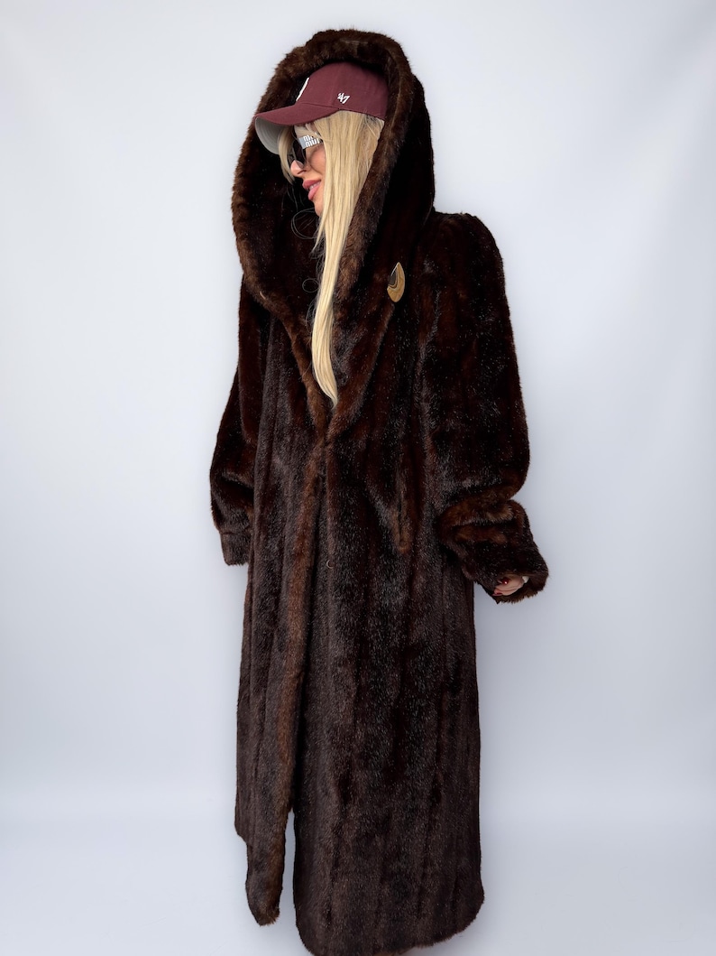 May include: A long, dark brown faux fur coat with a large hood. The coat has a button closure and a soft, plush texture. The hood is lined with a darker shade of brown. A maroon baseball cap is worn on the head.