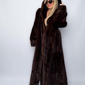 May include: A long, dark brown faux fur coat with a hood. The coat is floor-length and has a soft, textured appearance. The person is wearing a maroon baseball cap and sunglasses. The coat is a statement piece for cold weather.